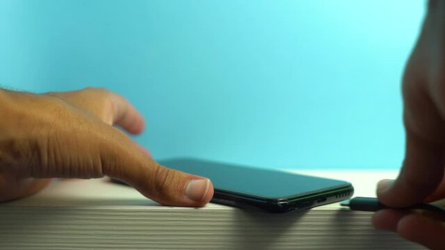 hand connecting charger to mobile. Mobile charging on top of white table with blue background. Young boy putting his phone to charge

