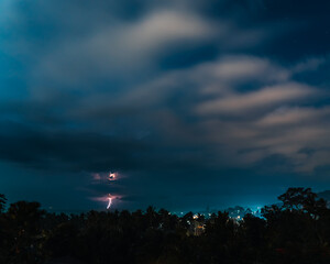 Lightnings, stars and dramatic clouds in the night sky over valley with trees, sea,  city and...