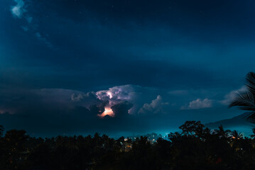 Lightnings, stars and dramatic clouds in the night sky over valley with trees, sea,  city and mountains. View from the top.