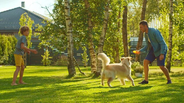 Handsome Father and Son Play Catch With Loyal Family Friend Golden Retriever Dog. Family Spending Time Together Training Dog. Sunny Day Idyllic Suburban Home Backyard. Slow Motion Shot