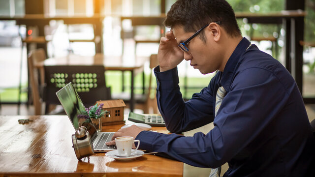 A handsome business man in a suit and glasses is working in front of a laptop, takes the handle and has an alarm clock and coffee mug, along with a house model placed next to a calculator.