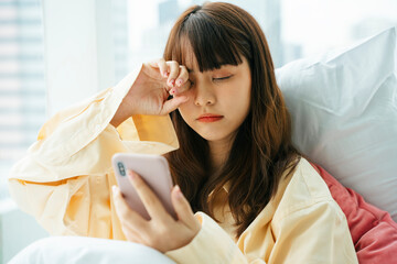 Sleepy asian dark hair woman wake up on a bed and use smartphone in the morning.