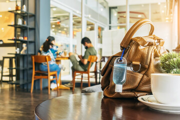 Mini alcohol gel bottle to kill Corona Virus(Covid-19) hang on a brown leather shoulder bag with hot coffee on table in cafe.New normal lifestyle.Health care concept.Selective focus on alcohol gel