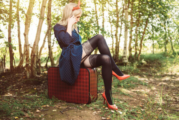 Sexy pinup girl in retro dress with red high heels and black stockings in the summer forest sitting on vintage suitcase	