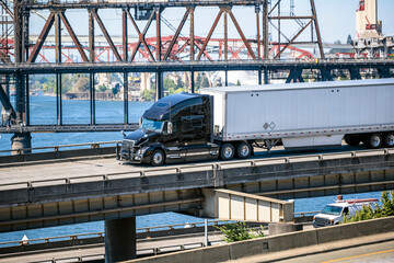 Naklejka premium Black big rig semi truck with dry van semi trailer transporting cargo running on the overpass road along the Willamette River in Portland
