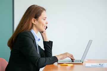 Happy pretty young businesswoman talking on mobile phone and smiling, working at computer in office, using laptop at table. Medium shot, side view. Communication or multitasking concept