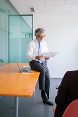 Focused Caucasian businessman sitting on table and reading document. Experienced senior bearded entrepreneur in eyeglasses meeting in conference room. Business and management concept