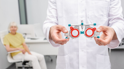 Doctor holding an optometric trial frame for eye exam in his hands. Senior woman sitting on...