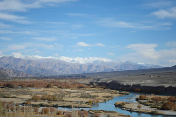A river runs through a valley in Ladakh, India, past some trees, shrubs and a few houses. The foothills of the Himalayas are in the distance, the higher mountains covered with snow. The sky is blue.
