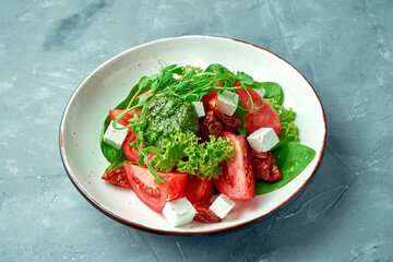 Healthy salad with sun-dried and fresh tomatoes, feta cheese and pesto in a white bowl on a gray background