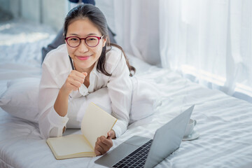 Young asian woman lying taking notes and working laptop computer on bed at home.