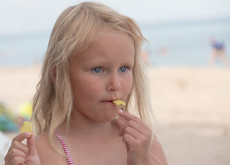 A little five-year-old blonde with chips in her mouth is thinking about something.