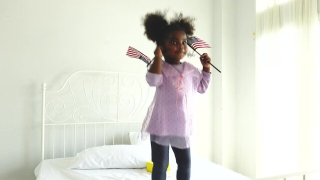Happy cute active little african american kid girl holding american flag on bed
