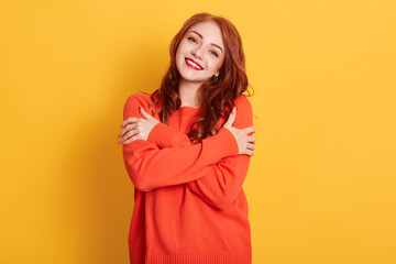 Cheerful young romantic woman expresses self love and care, smiles gently, wears orange oversized jumper, embraces own body, standing against yellow background.