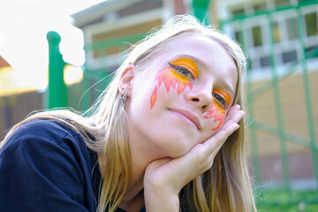 Beautiful teenager girl with face painting lying on a lawn. smiling blonde girl with fire and flame festive face painting. fire face art on teenager girl's eyes and cheeks. 