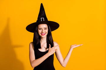 Photo of cute pretty young magician lady toothy smile hold empty space directing finger make perfect recommendation wear black wizard headwear dress isolated bright yellow color background