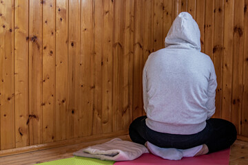Fototapeta na wymiar A girl in a light sweater with a hood on her head sits in meditation on the floor and looks at the corner of the walls.