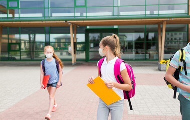 education, childhood and pandemic concept - group of elementary school students wearing face protective medical mask for protection from virus disease with backpacks walking and talking outdoors