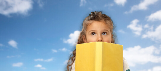 reading, education and childhood concept - little girl hiding behind yellow book over blue sky and clouds in background
