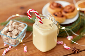 christmas and seasonal drinks concept - eggnog in glass mug with candy cane decoration, cinnamon buns, fir tree brunch and garland lights on wooden background