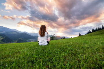 Obraz na płótnie Canvas Girl in a white shirt with red hair sitting on the lawn in the mountains. Green grass and sunset in the Ukrainian Carpathians.