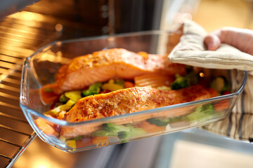 food cooking, culinary and people concept - young woman with potholder taking baking dish with salmon fish and vegetables out of oven at home kitchen