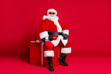 Full length photo of retired old man white beard sit big giftbox hold dumbbells warming up arms wear x-mas santa costume glove coat belt sunglass cap boot isolated red color background
