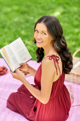 leisure and people concept - happy smiling woman sitting on picnic blanket and reading book at summer park