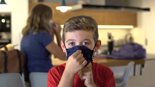 Caucasian boy puts on a protective mask at home looking at camera on First day School. A girl behind puts on a mask too. Concept new rules, new normality during Coronavirus, COVID-19.