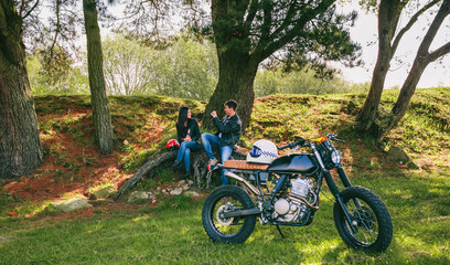 Fototapeta premium Young couple resting from a motorcycle trip taking picture outdoors with motorbike in foreground. Selective focus on couple in the background