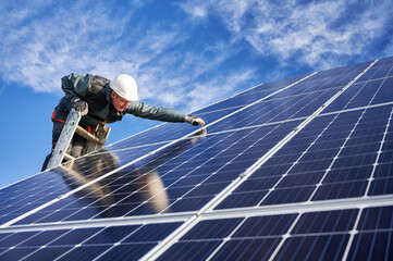 Male technician in white safety helmet standing on ladder and installing photovoltaic solar panel...