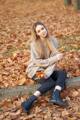 Autumn portrait. Blonde pretty girl with beautiful long hair wearing youth cosy sweater and black jeans holding a bouquet of maple leaves in the hand.
