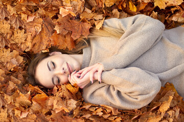 Happy smiling cute girl with closed eyes liying in the autumn leaves and enjoying it, holding face...