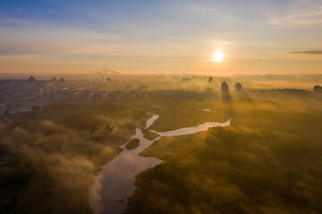 The Svisloch River in Minsk and the Rising Sun, through the fog in the city! Loshitskiy Park. In the foreground is a small village and a river, in the distance the skyscrapers of the city.