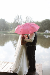 Bride and groom standing under pink umbrella under summer rain hugging and holding hands. For better or worse. Wedding promises and vows idea. 