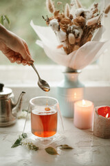 Brewing tea. Double wall glass cup with black tea on old vintage window. Hand with spoon makes tea dropping. Flowers in vase and candles in the background.