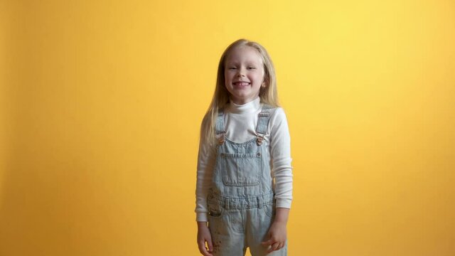 The blonde girl laughs and points a finger to the camera. Contempt, ridicule concept. Yellow background. Girl in denim overalls casual wear.