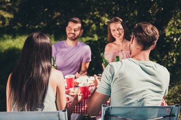 Back rear spine view photo of four fellows girls guys sit table enjoy summer vacation have fun speak talk eat lunch drink beer bottle under green leaves tree garden outdoors