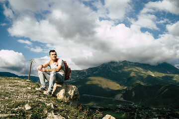 Young man sitting on hill. Dreamy male sitting on altitude and looking at Peaks of magnificent rocks located against bright cloudy sky