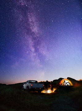 Friends near Bonfire, Pickup Truck, Tent and Bike in the Mountains under Night Sky with Milky Way. Adventure and Travel
