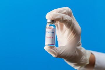 A hand in a medical glove holds a bottle of COVID-19 vaccine on a blue background.