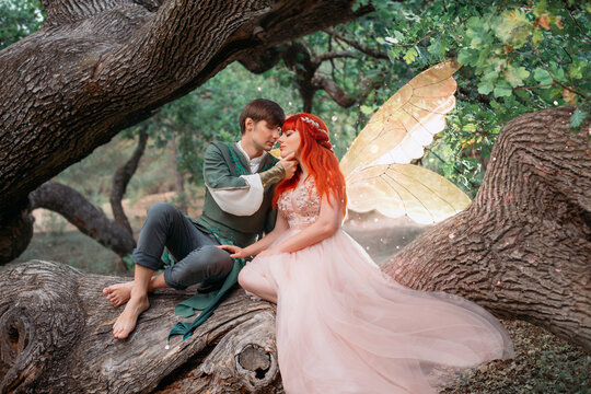 Young couple in love. A man and a woman are hugging on a huge tree. Themed creative wedding bright fantasy photography. Fairy woman in long pink dress with bright golden wings. A man in an elf costume
