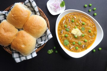 Indian Mumbai Street style Pav Bhaji, garnished with peas, raw onions, coriander and Butter. Spicy thick curry made of out mixed vegetables, served with paav over black background with copy space. 