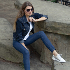 A stylish blonde in a denim jumpsuit sits on a rock in a city park. A girl in jeans and a white t-shirt at a stone table