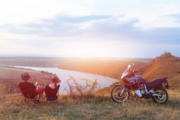 A couple of tourists, a man and a woman, enjoy the beautiful view. They sit on folding chairs. Motorcyclist, bikers vacation. Tourism and camping. Sunset. space for text. Soft light.
