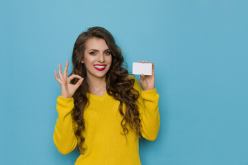 Smiling Woman In Yellow Sweater Holds White Card And Shows Ok Hand Sign