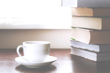 A white cup of coffee stands on a wood table next to the books