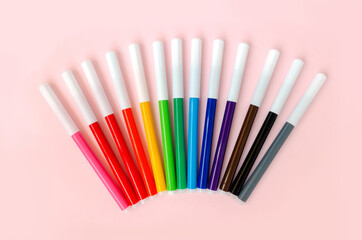 Markers are fanned out according to the colors of the rainbow on a pink background. Top shot of school supplies.