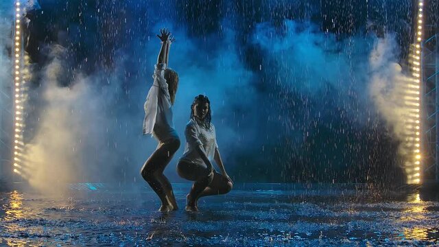 Two young women passionately dancing salsa elements in raindrops and smoke. Female silhouettes in wet bodysuits move erotically in slow motion.