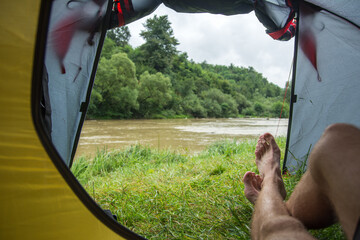 Tent by the river. Gloomy sky and fog in the distance. Dirty brown water. Trees in the valley. Green grass. Leisure and travel. Camping. Motivation. Inside the tent.